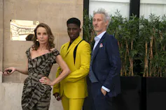 A woman and two men stand outside an office. The woman wears a chic black dress, and the men wear suits, one bright yellow, one navy.