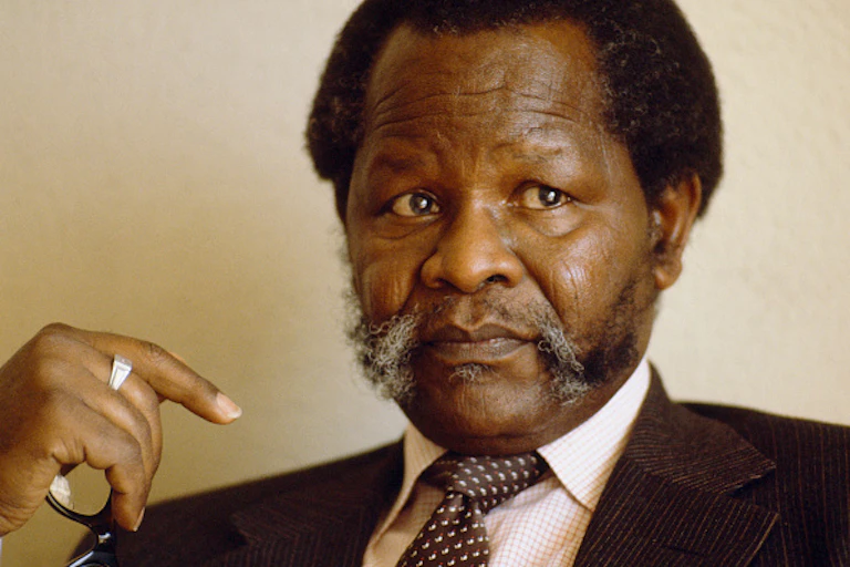 A serious looking man with a ring on his middle finger, wearing shirt, tie and corduroy jacket holds specs in the palm of his hand.