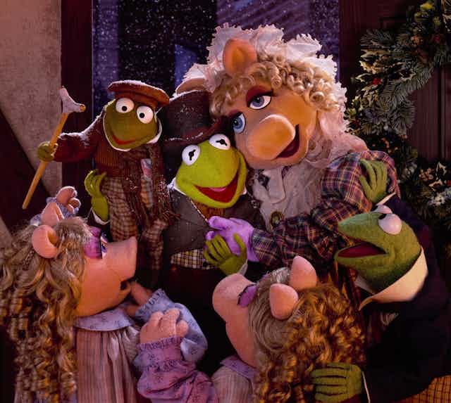Miss Piggy, Kermit and their children, two frogs and two piglets, embrace. They all wear Dickensian clothing.