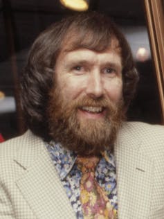 Jim Henson has long hair and a bushy beard. He wears 1970s attire, including an orange tie and cream blazer jacket.