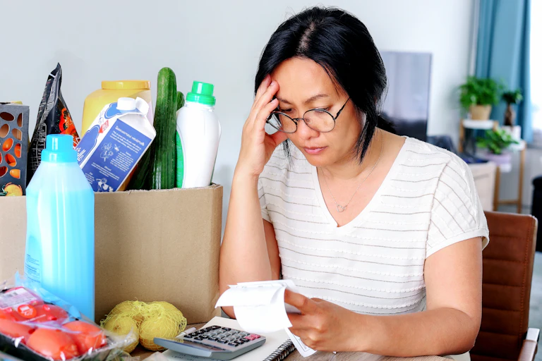 A woman holding receipts from supermarket with calculator, groceries in the background, finances, stress, worry.
