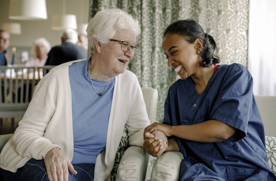 Smiling and laughing female nurse holding the hand of a senior woman sitting at retirement home, with other seniors in the background.