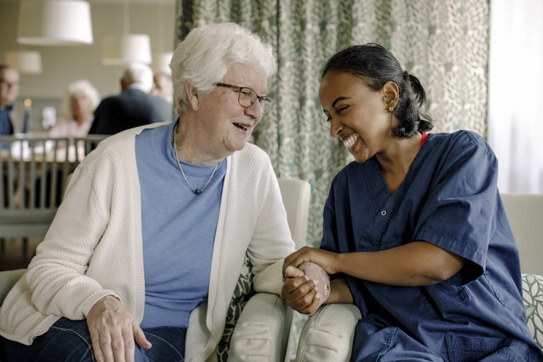 Smiling and laughing female nurse holding the hand of a senior woman sitting at retirement home, with other seniors in the background.