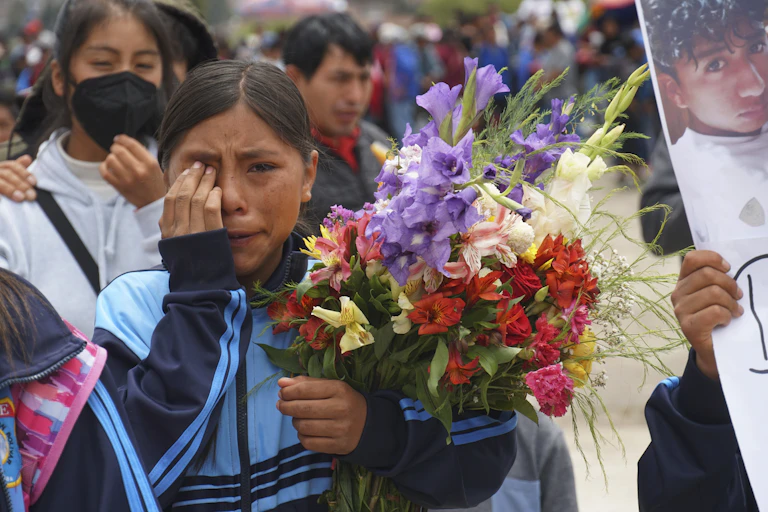 A dark-haired girl carrying flowers weeps. Someone next to her carries a sign featuring a boy's face.