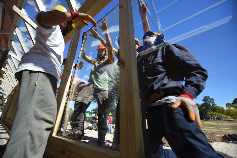 A group of people wearing hard hats install a large window.