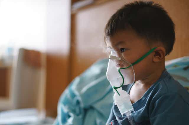 A sick 3-year-old boy sits in his hospital bed wearing a nebulizer that fits over the nose.