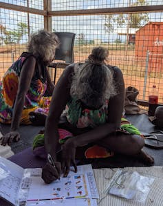 A First Nations person, Rarrapul sits with others on the floor and writes in a folder.