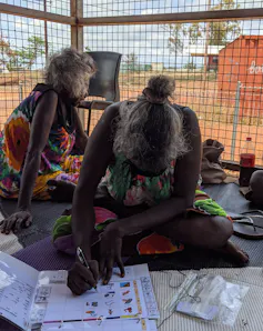 A First Nations person, Rarrapul sits with others on the floor and writes in a folder.