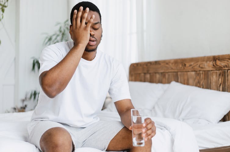 A young man with a hangover sits in bed holding a glass of water. One hand holds his head in pain.