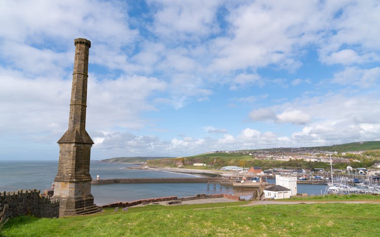 A stone monolith stands on a hill overlooking a tranquil harbour.