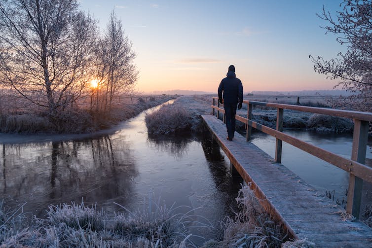 Man walking over a small footbridge in the frozen countryside during a winter sunrise.