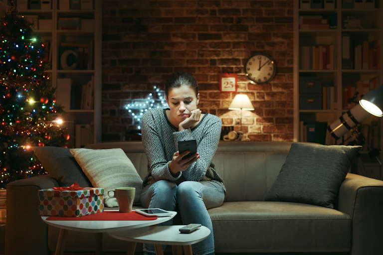 Woman celebrating Christmas at home alone: she is sitting on the sofa and waiting for messages on her smartphone