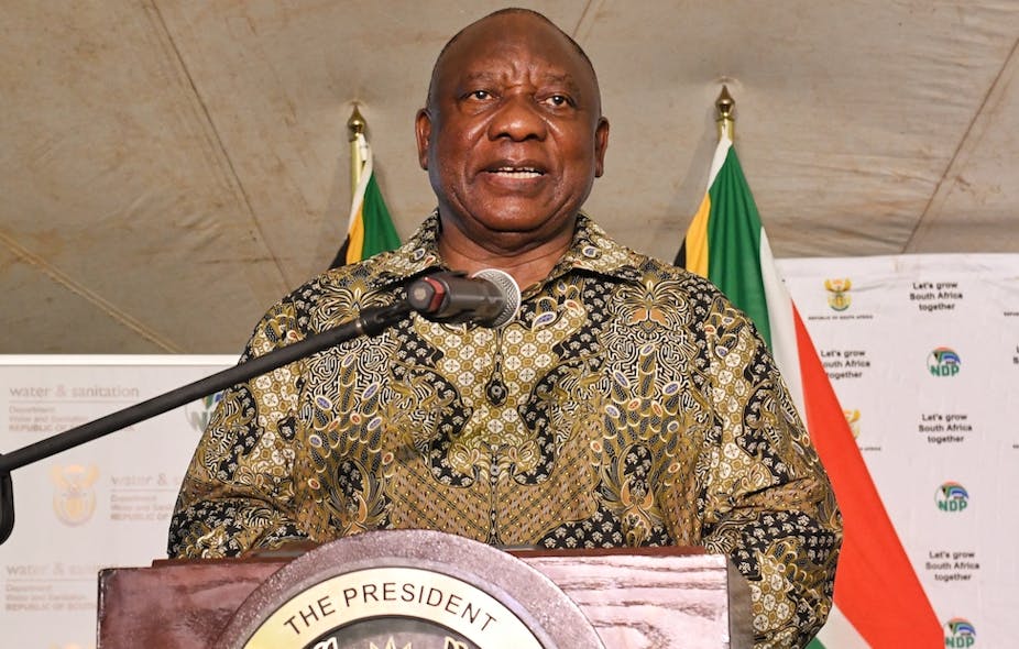 A man speaks at a podium with his back to two South African flags.
