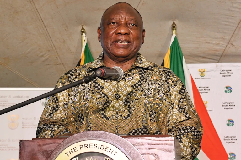 A man speaks at a podium with his back to two South African flags.