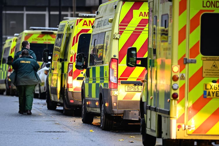 Paramedics in green walk past a row of ambulances with lights on.