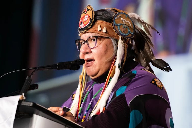 A woman in an Indigenous headdress speaks into a microphone.