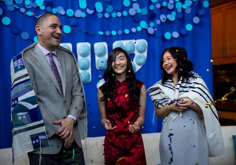 A man and woman flank a teenage girl in a red and black dress as they all smile and laugh.