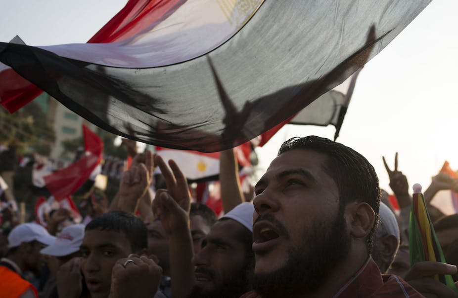 A man looks out from underneath an Egyptian flag with demonstrators behind him.