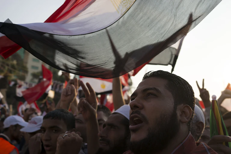 A man looks out from underneath an Egyptian flag with demonstrators behind him.