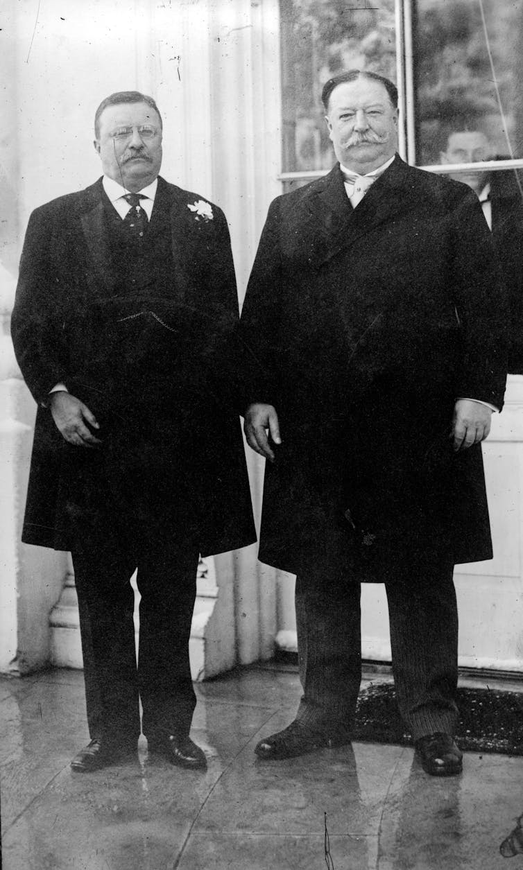 Two white men in business suits and overcoats stand next to each other.