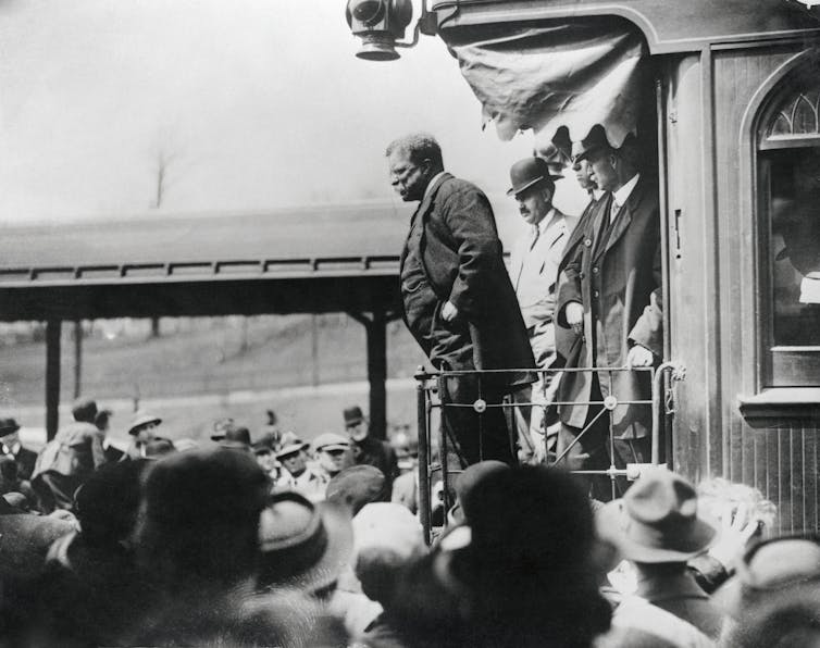 A middle-aged man dressed in a business suit stands above a crowd of people as he delivers a speech.