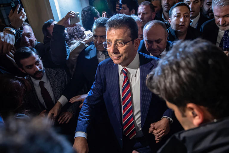 A man in glasses and a blue striped suit walks through a crowd.