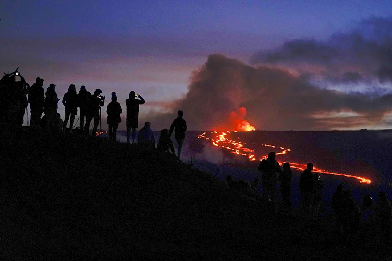 A group of people taking photos stand silhouetted against a volcano erupting in the distance. A massive cloud of smoke billows from the volcano against the sky.