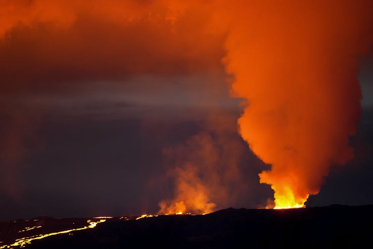 A column of orange smoke billows from the mouth of a volcano. A stream of yellow-orange lava flows down the side of it.