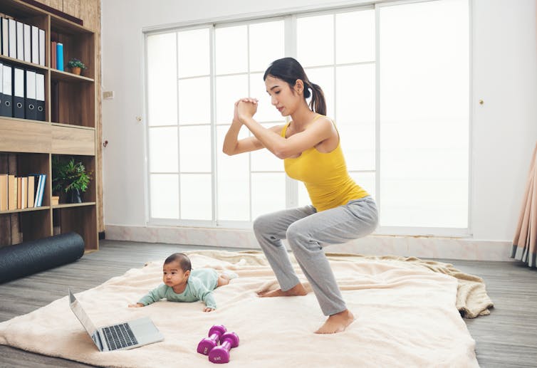 Woman in yellow sportswear exercising with a baby on the floor as well as a pair of dumbbells and a laptop