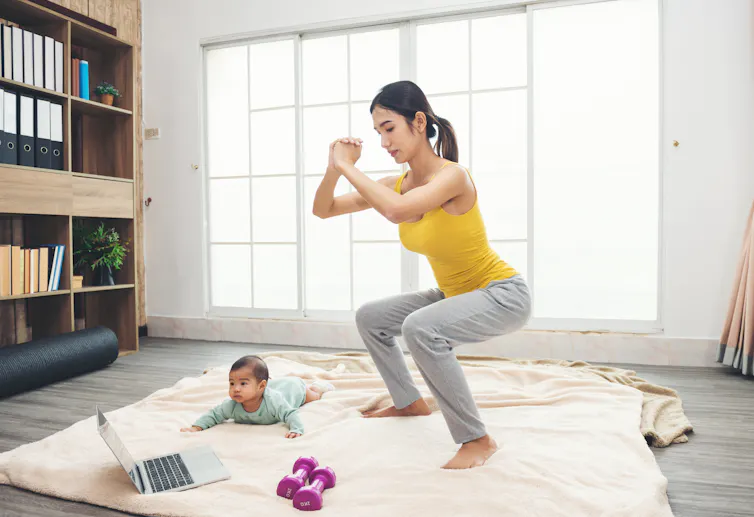 Woman in yellow sportswear exercising with a baby on the floor as well as a pair of dumbbells and a laptop