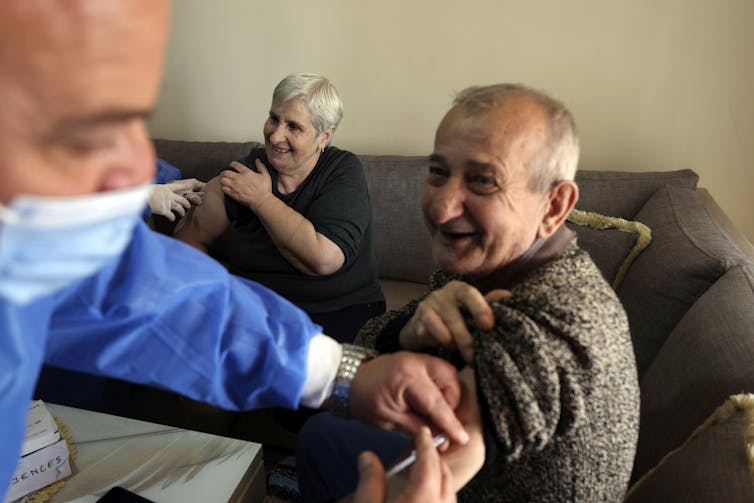 A smiling grey-haired man and woman get vaccinated by someone in a mask and blue scrubs.