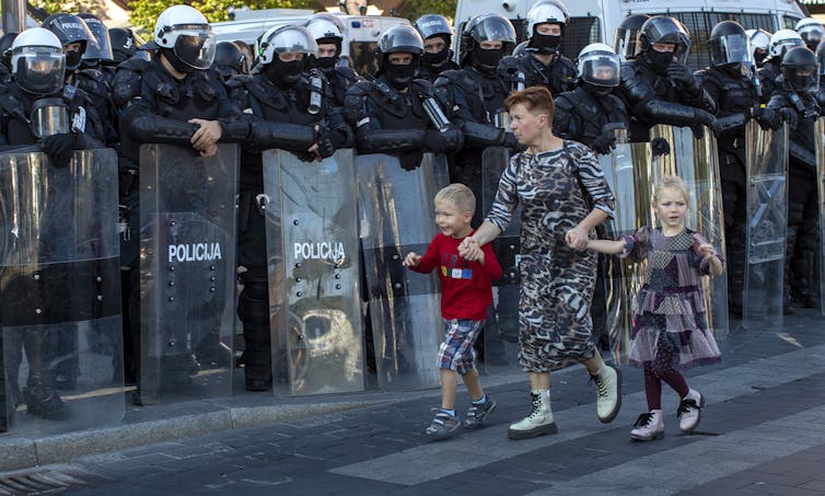 A dark-haired woman in an animal-print dress walks past a line of riot police holding the hands of two blonde children.
