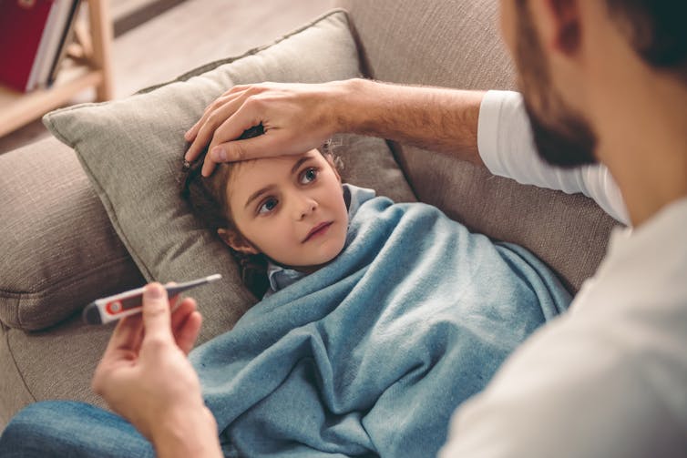 A man feels his daughter's forehead, holding a thermometer.