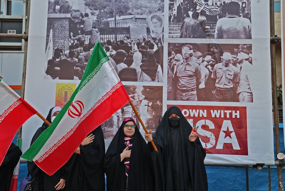 Femmes voilées avec drapeaux de l'Iran devant un montage de photos de la prise d'otages de l'ambassade américaine à Téhéran en 1979