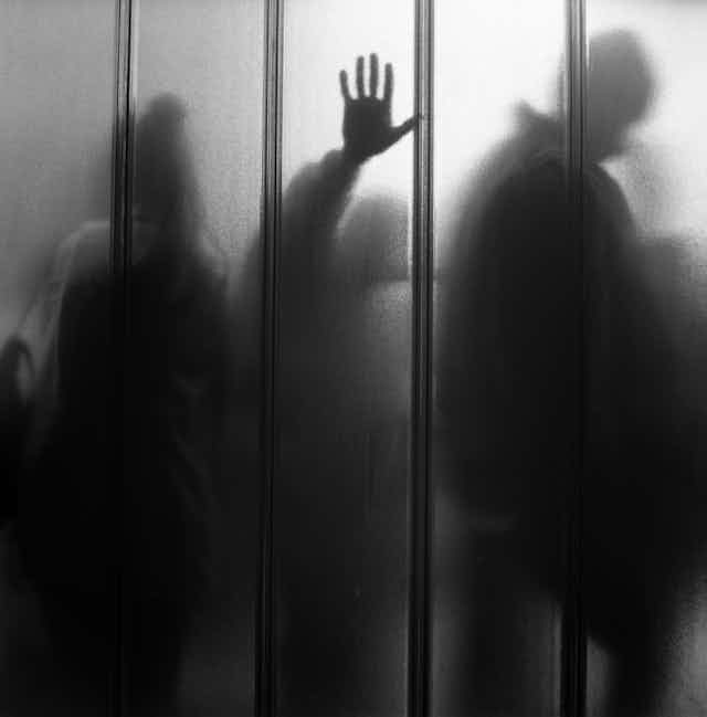 Black and white photo of a translucent wall with people queuing on the other side, one person has their hand pressed up against the glass