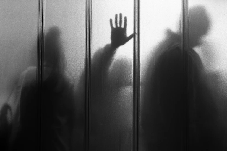 Black and white photo of a translucent wall with people queuing on the other side, one person has their hand pressed up against the glass