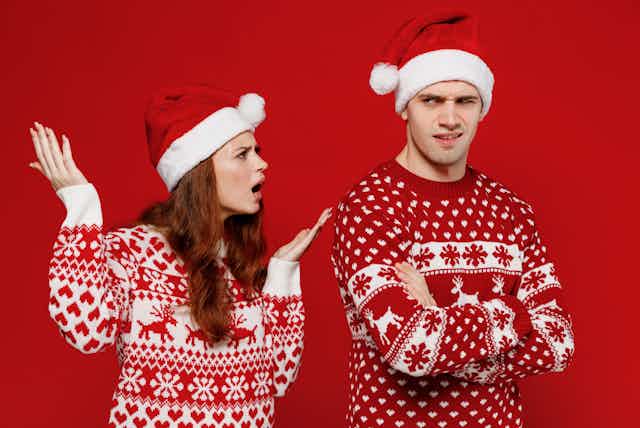 A young man and woman in red Christmas jumpers and santa hats, arguing with each other