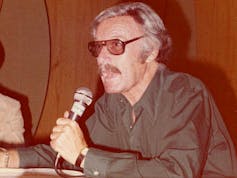 Stan Lee wears large tinted glasses, wears a green shirt and holds a microphone. His hair is grey.