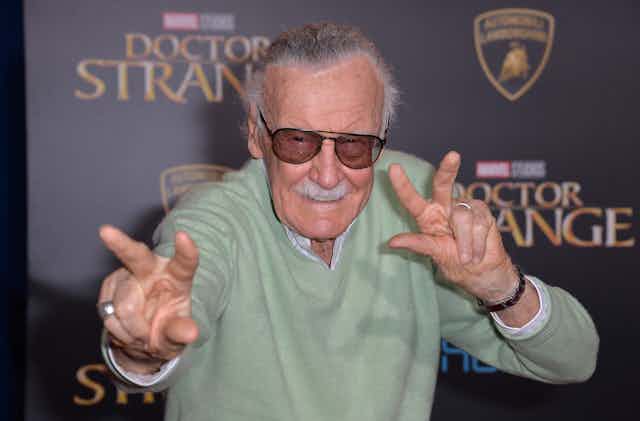 Stan Lee poses in dark sunglasses imitating Doctor Strange's signature hand movements. He wears a sage green jumper. 