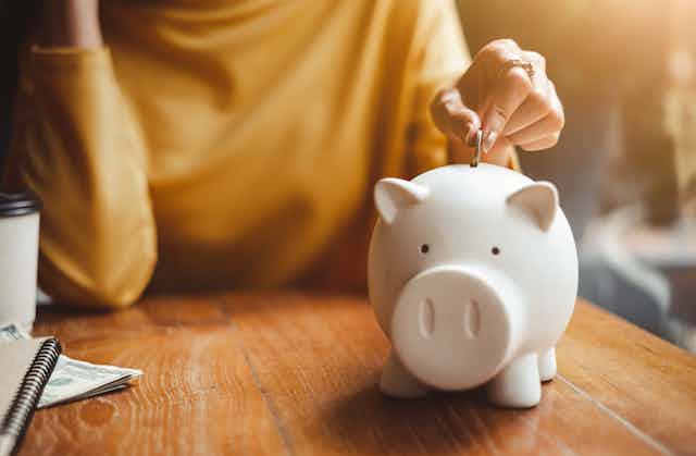 A woman wearing yellow shirt and leaning on table puts a coin in a white piggy bank