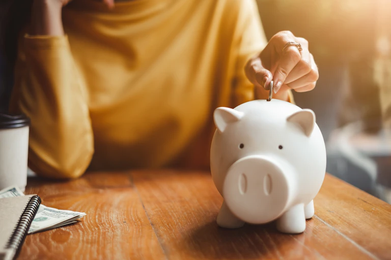 A woman wearing yellow shirt and leaning on table puts a coin in a white piggy bank