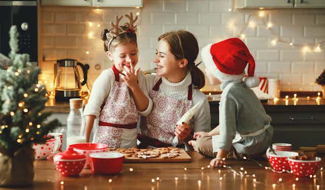 A woman ices holidays cookies with her two children in a kitchen. One child wears a reindeer headband while the other wears a Santa hat.