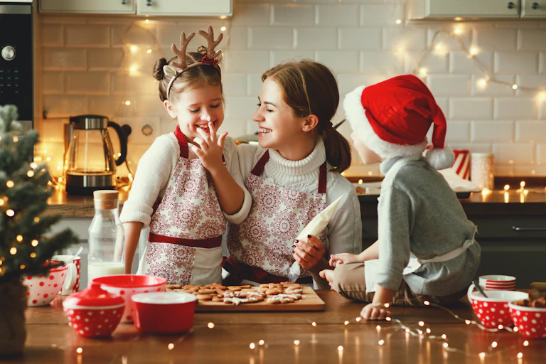 A woman ices holidays cookies with her two children in a kitchen. One child wears a reindeer headband while the other wears a Santa hat.