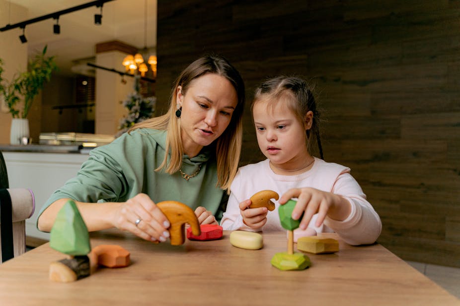 A woman sits at a table with a girl with Down Syndrome playing with toys
