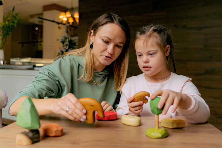 A woman sits at a table with a girl with Down Syndrome playing with toys