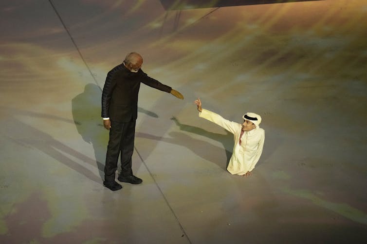 A man wearing black holds out to his arm to a smaller man wearing Arabic clothing who is also holding out his arm.