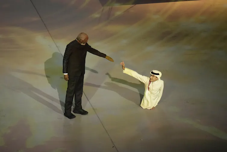 A man wearing black holds out to his arm to a smaller man wearing Arabic clothing who is also holding out his arm.