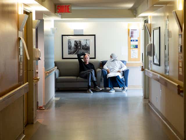 A man and a person in PPE are seen sitting on a sofa at the end of a corridor
