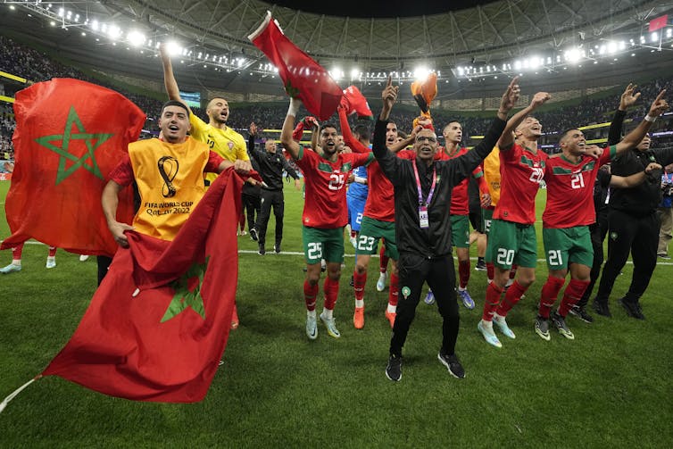 Moroccan soccer players wearing red and green celebrate on a football pitch with Moroccan flags.