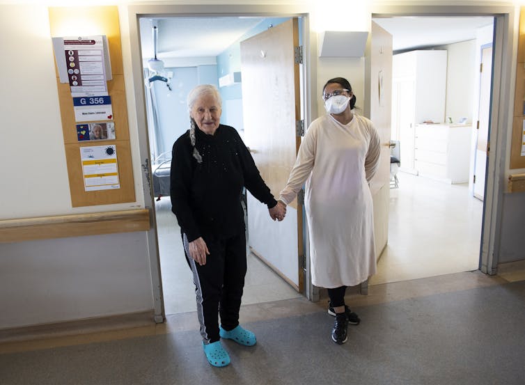 A woman with gray hair and a woman in scrubs and a face mask hold hands in a corridor